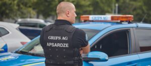 Apsardzes darbinieks formas tērpā stāv blakus zilai apsardzes firmas mašīnai
